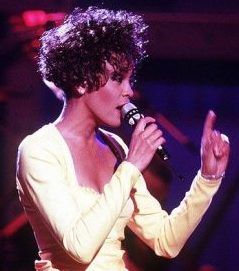 Whitney Houston on stage singing into a microphone. She has short, dark brown curly hair, and she's wearing a yellow pant suit.
